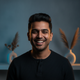 Headshot of a smiling South Asian man in his late 20s, with a confident expression, set against a backdrop of slate blue and 